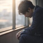窓際でうつむきながら悩む男性の様子(労災申請・メンタル不調のイメージ)